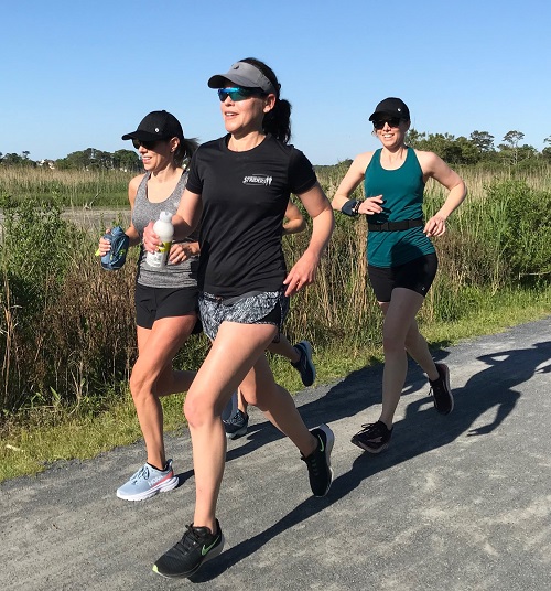 Alex Barrage running with two other women on a crushed gravel trail through marshy land on a sunny day