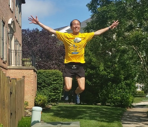 Kam Yee, wearing a yellow tech shirt and black shorts, jumping very very high in front of a brick row house