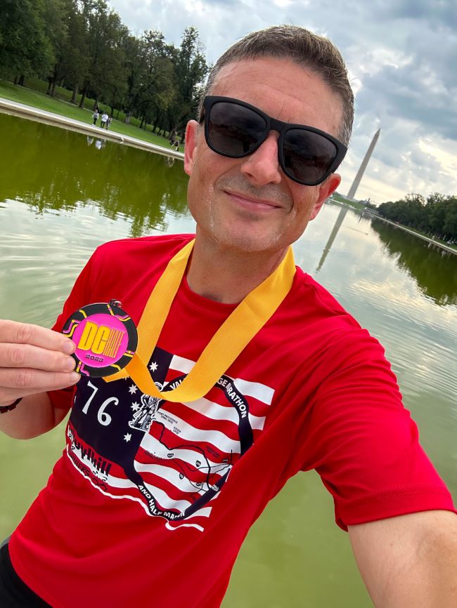 Robert Kelley holding his DC Half finisher medal while standing in front of the Reflecting Pool on the National Mall