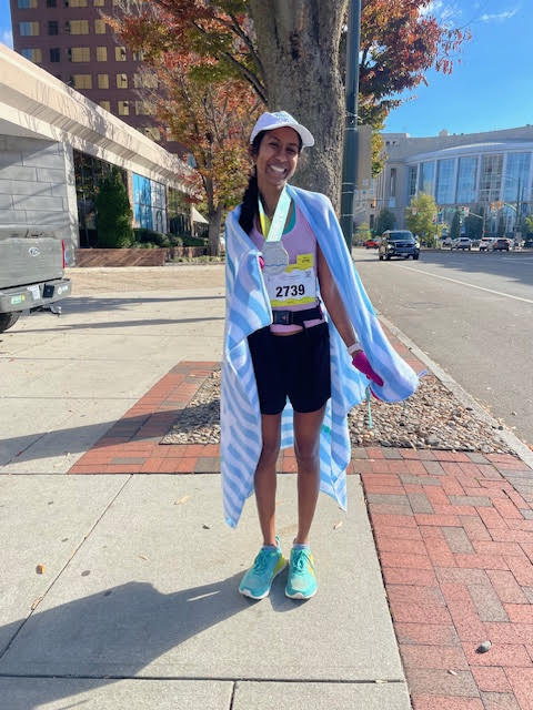 Vinona Muralidaran standing on a city sidewalk holding a finisher medal while being draped in what looks like a blue and white striped beach towel