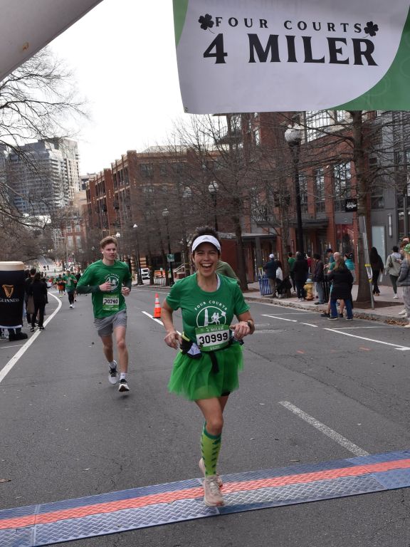 Kristina Graff crossing the finish line of the 2026 Four Courts Four Miler while wearing the green race shirt and and a celebratory green tutu