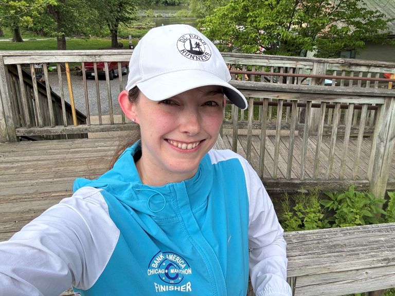 Rae Bimmerle smiling while taking a selfie at Fletchers Cove in Washington, DC. She is wearing a white DC Road Runners hat and a blue Chicago Marathon finisher's jacket