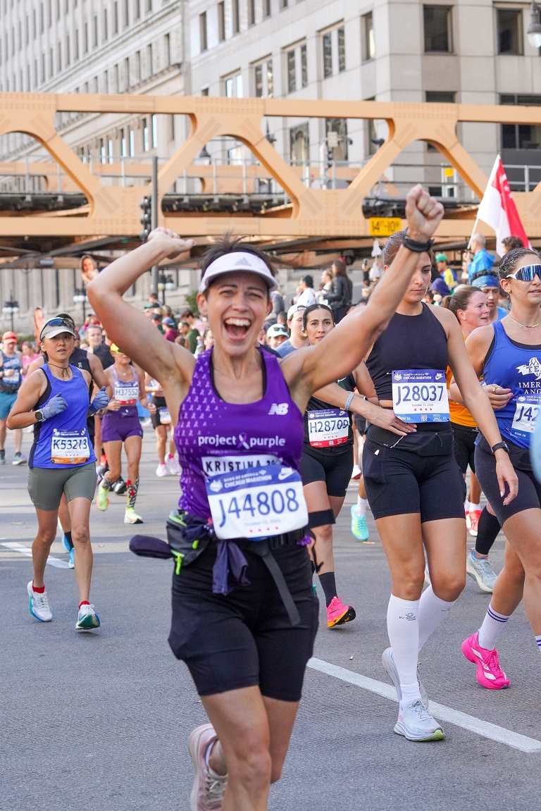 Kristina Graff running for Project Purple in the Chicago Marathon. She is smiling and waving her hands in the air while running in a crowd